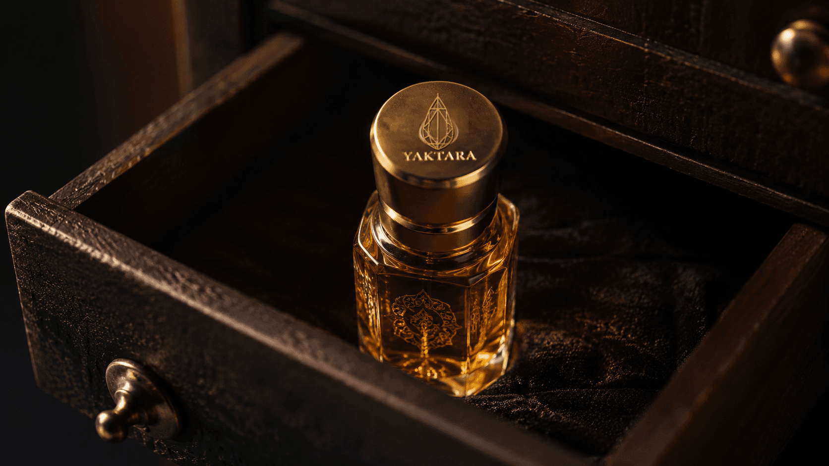 A Yaktara attar bottle with gold cap engraved with the drop-mark logo, standing in an open velvet-lined drawer lit by warm golden light.